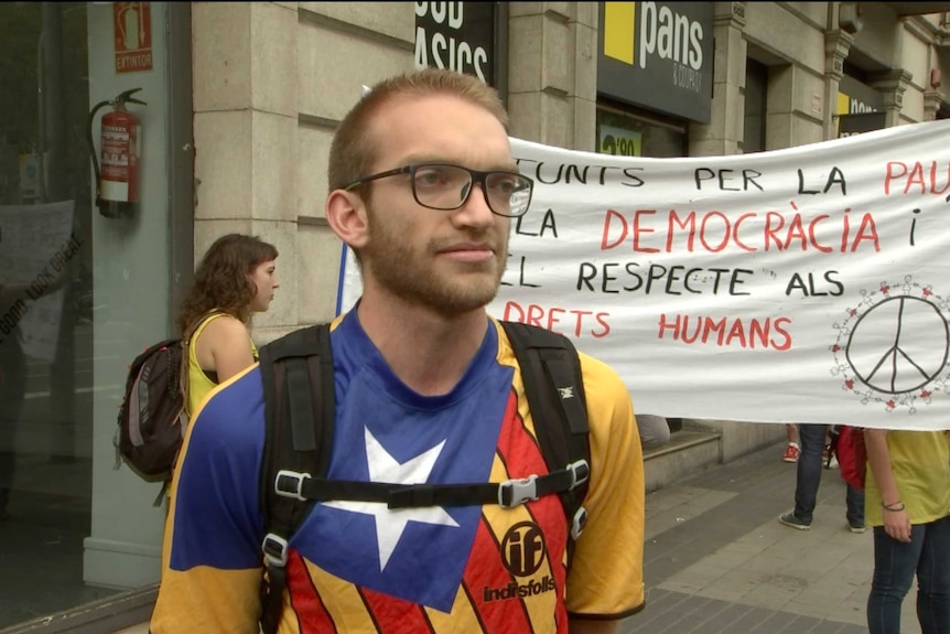 A young man stands in a street in Barcelona in front of a banner calling for democracy.