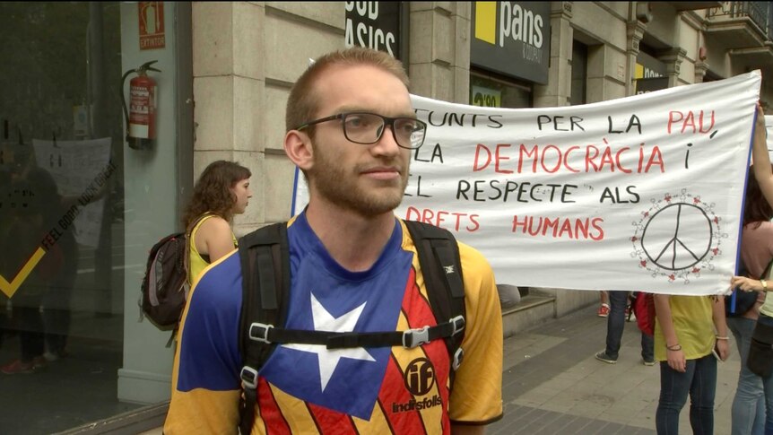 A young man stands in a street in Barcelona in front of a banner calling for democracy.