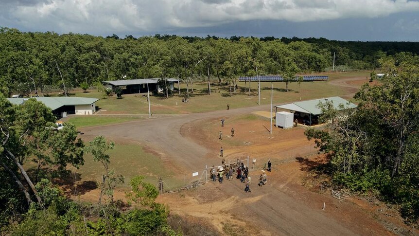 An aerial image of two groups gathered on opposing sides of a cyclone fence, in a compound in the bush