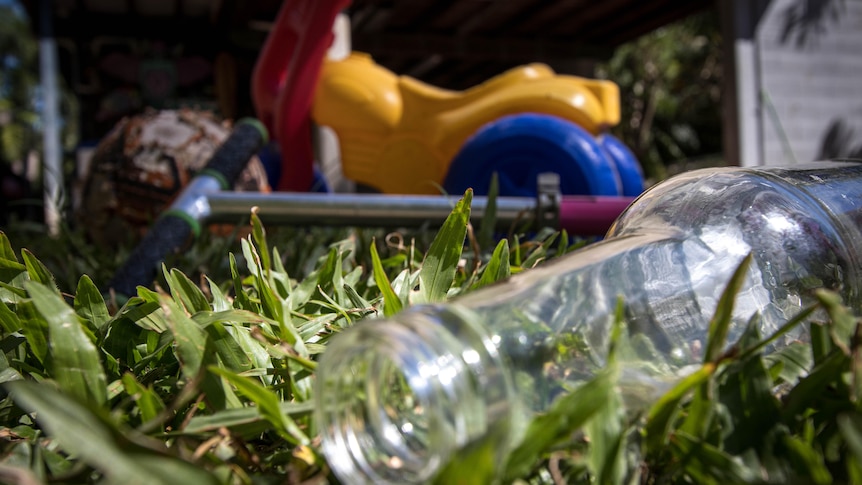 A bottle of beer lies empty in a yard, with kids toys in the background.