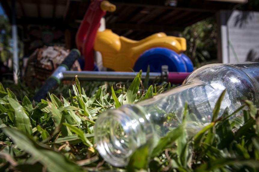 A bottle of beer lies empty in a yard, with kids toys in the background.