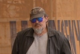 A man with a beard wearing a brown jacket, brown cap and sunglasses