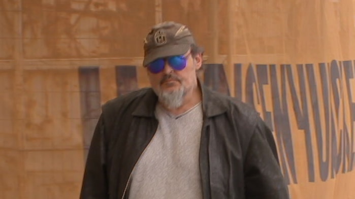 A man with a beard wearing a brown jacket, brown cap and sunglasses
