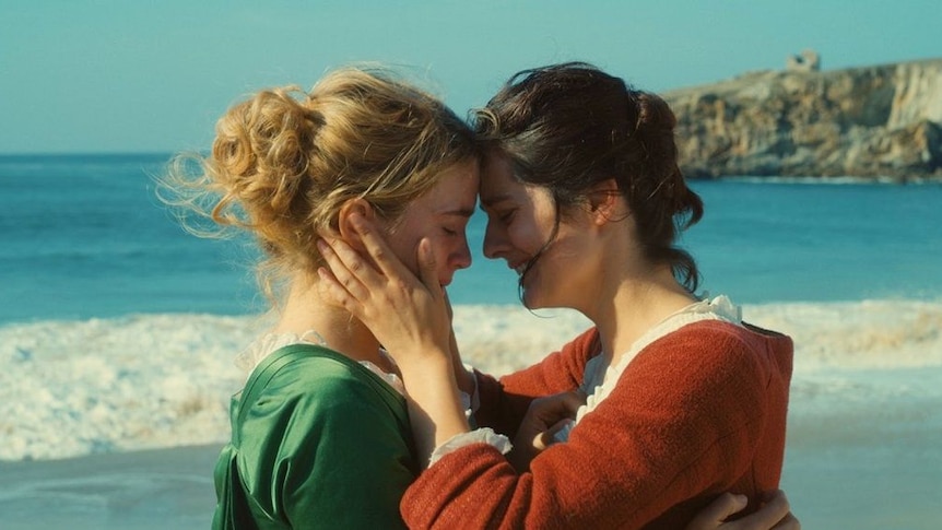 Two women embrace with foreheads touching, crying, in front of the ocean, dressed in 18th century clothes