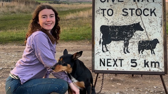 A young woman kneels next to her Kelpie dog, with a 'give way to stock' sign and sunset in the background.