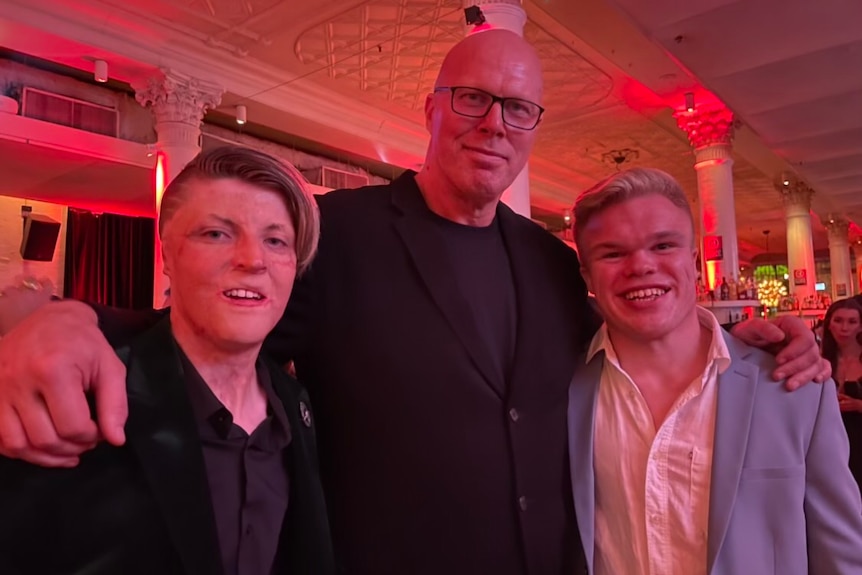 Three men in suits take a selfie on the red carpet, and the room is lit in red and pink.