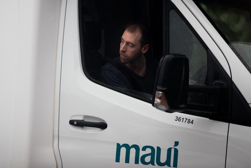 A man sticks his head out of the window of a white vehicle with the word Maui written on its door and looks backwards.