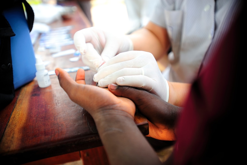 A young African student has their hand on a wooden table while a nurse takes a blood sample