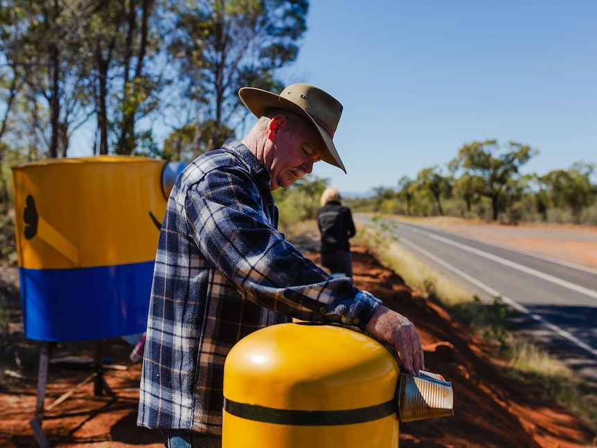 A man wearing an Akubra hat, inspects his hand-made Minions. Capricorn Highway and bushland in background.