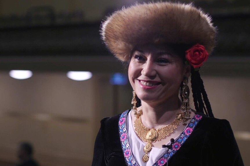 A woman with a fur-lined hat and rose in her hair smiles. She wears gold jewellery and intricate patterned cloak