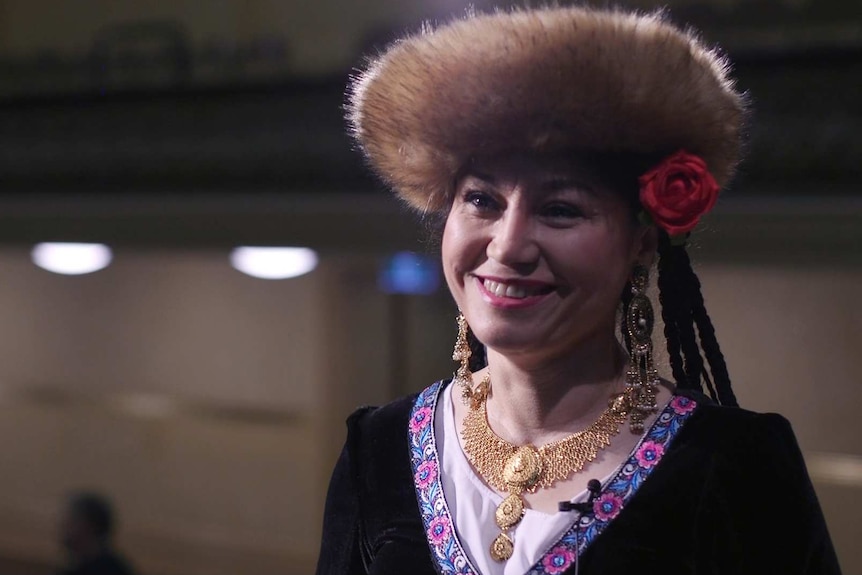 A woman with a fur-lined hat and rose in her hair smiles. She wears gold jewellery and intricate patterned cloak