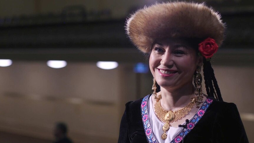 A woman with a fur-lined hat and rose in her hair smiles. She wears gold jewellery and intricate patterned cloak