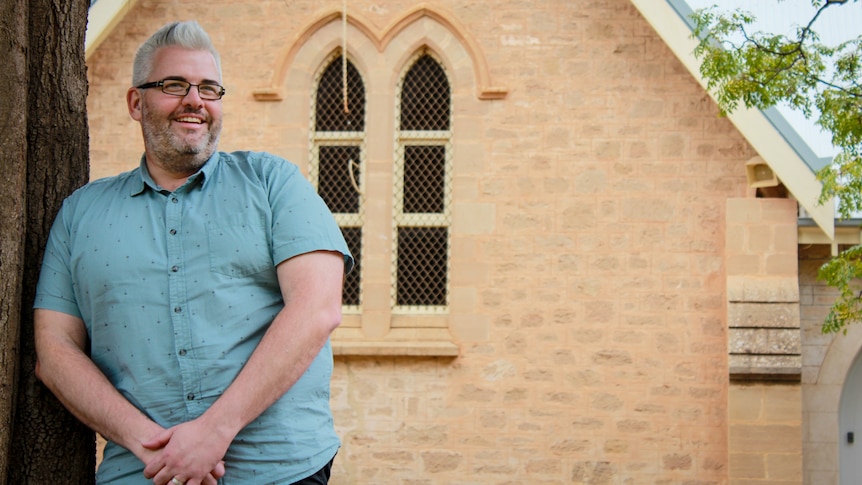 A man with silver hair, a beard and glasses leaning against a tree outside a sandstone church.