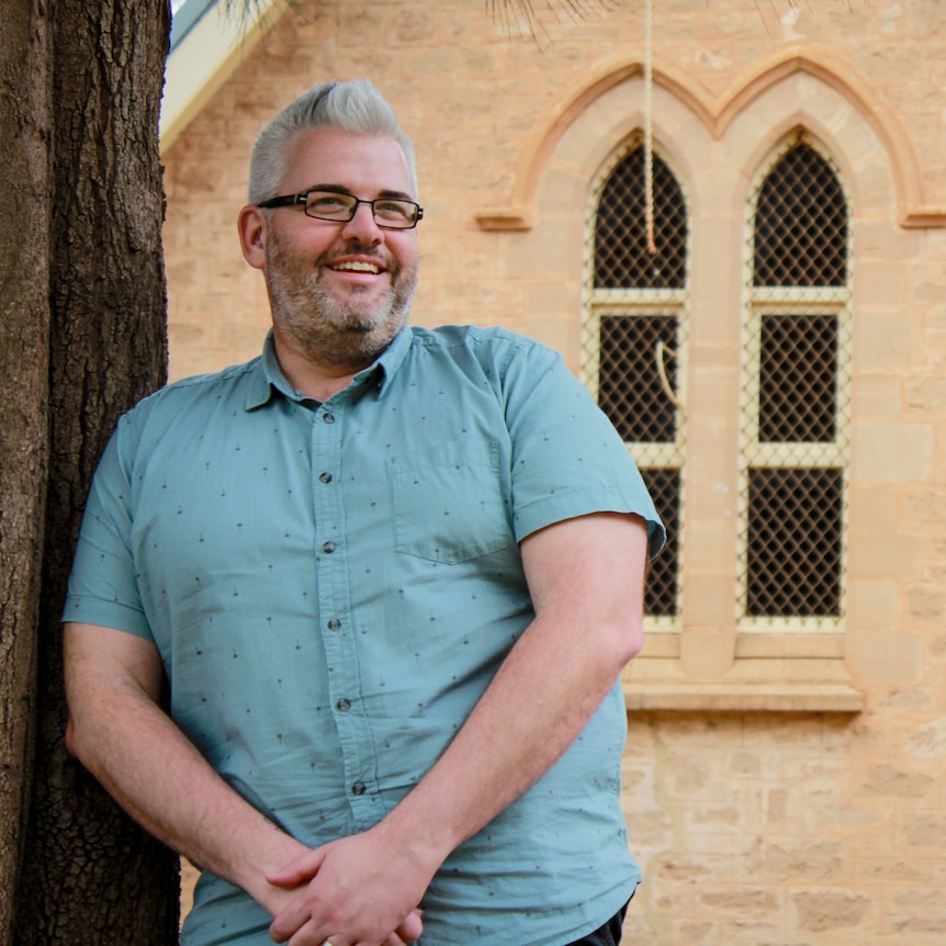 A man with silver hair, a beard and glasses leaning against a tree outside a sandstone church.