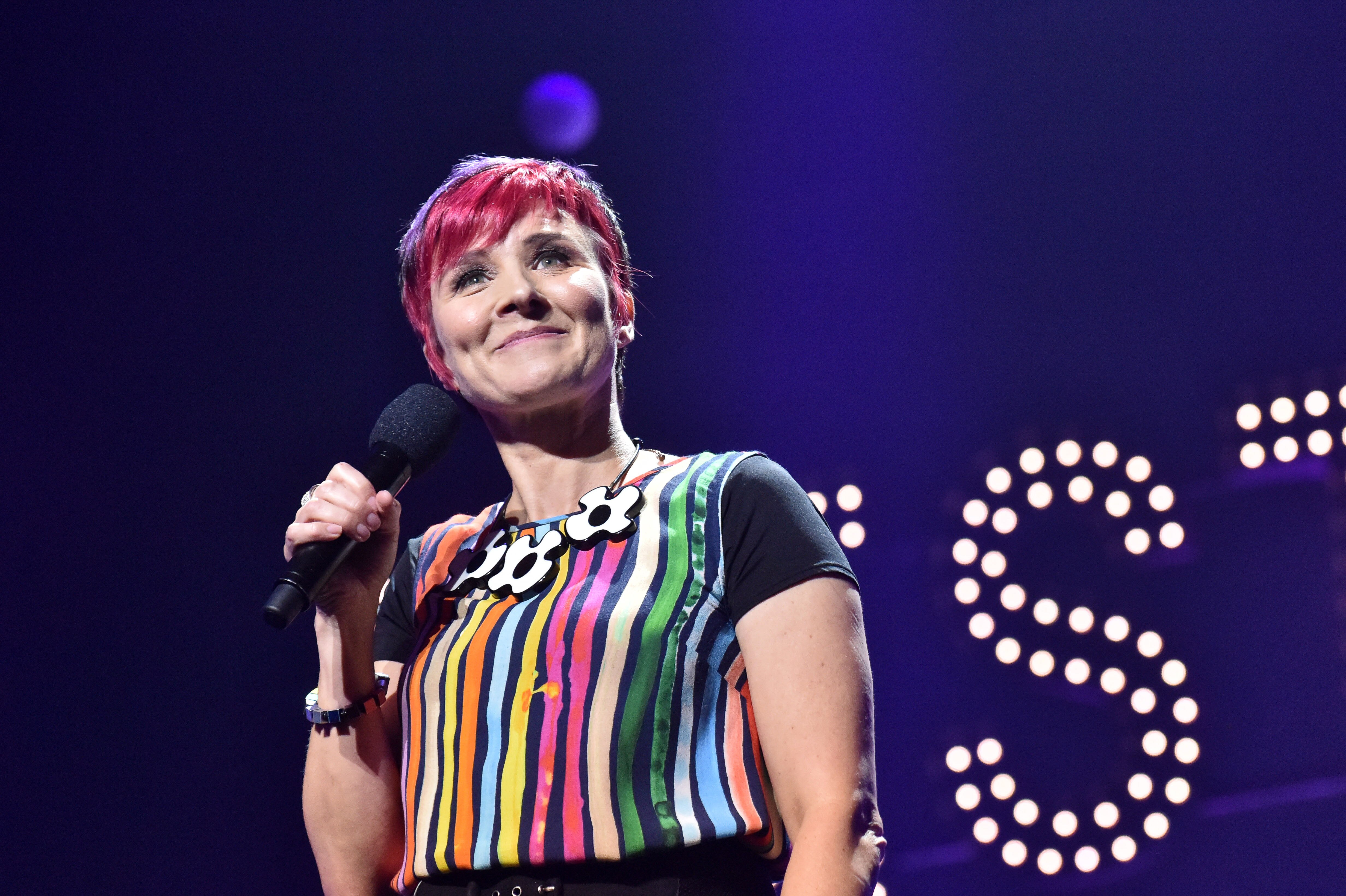 Cal Wilson smiling with a microphone wearing brightly-coloured clothing.