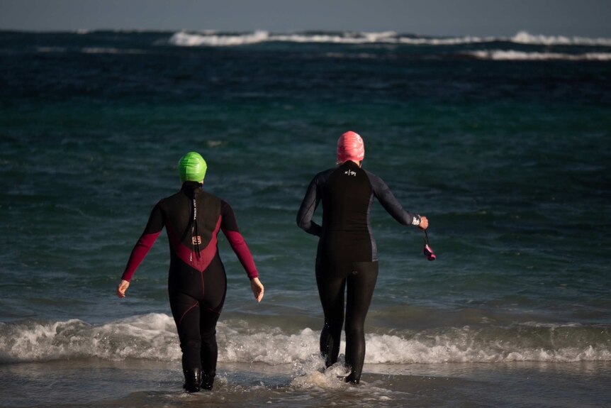 two women in wetsuits walk into the ocean