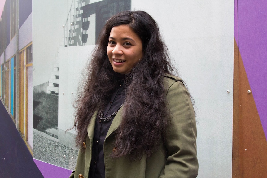 Colour photography of Muslim Zainab Kadhim standing in front of grey and purple wall.