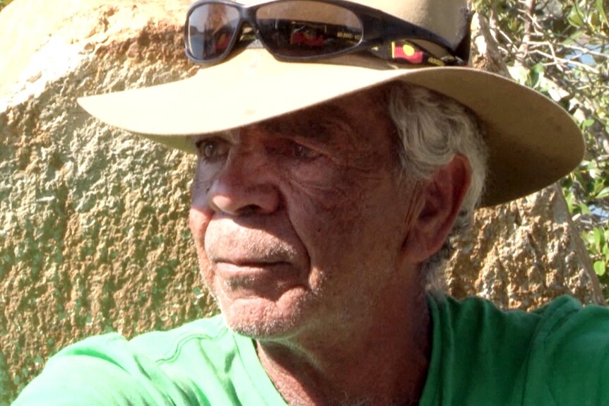 Aboriginal elder in an Akubra hat with sunies on top sitting in front of a rock looking away into the distance.