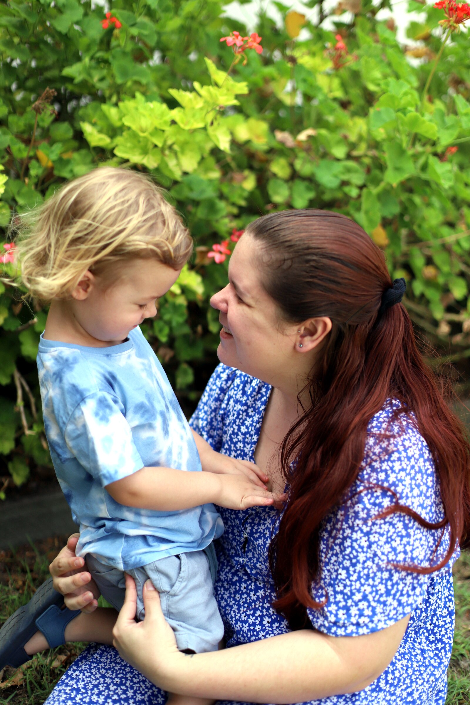 Tasha Torzsa looks at her son while smiling. They wear blue and kneel in front of a green shrub with pink flowers.