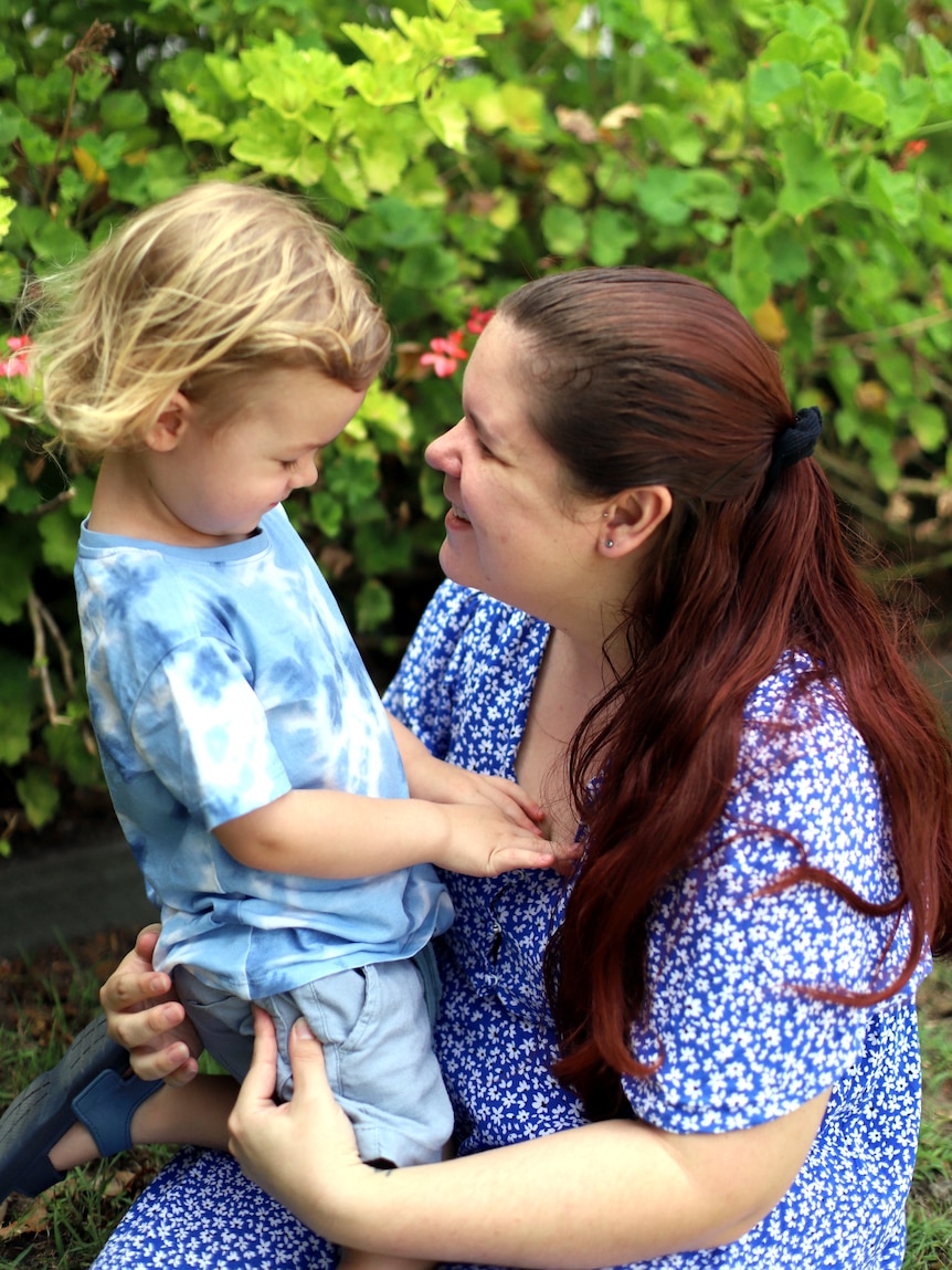 Tasha Torzsa looks at her son while smiling. They wear blue and kneel in front of a green shrub with pink flowers.