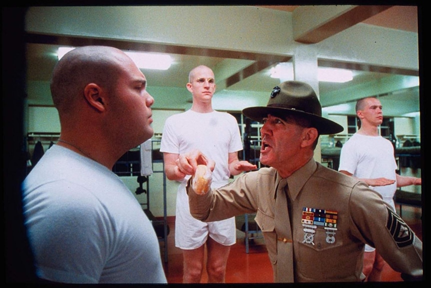 Still from Full Metal Jacket showing Ermey as the Gunny holding a bread role and berating a male recruit in a dormitory
