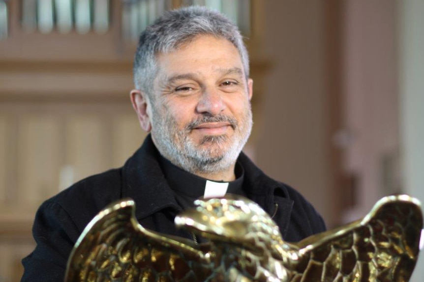 Father Peter Manuel smiles posing for a picture with a golden eagle ornament in front of him.