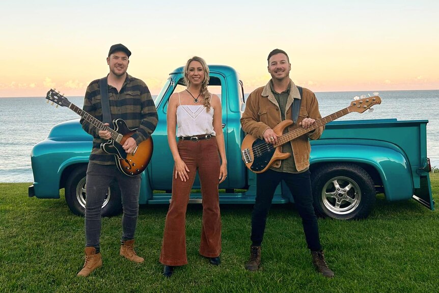 Two men with guitars and a women in the middle standing in front of a vintage car.