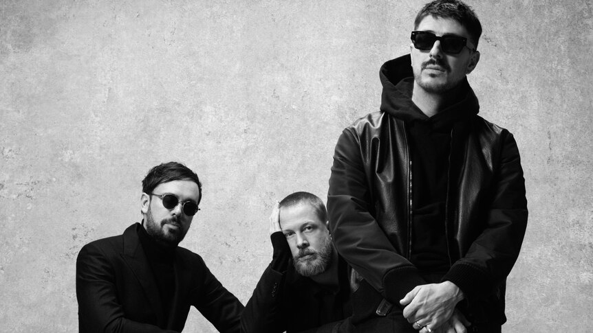 Black & white photo of Mumford & Sons styeld with sunglasses, wearing black at a table 