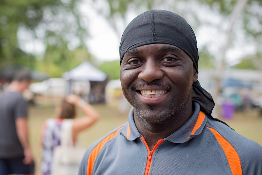 A man of African appearance wearing a fabric skull cap smiles for a portrait photograph at a market day.