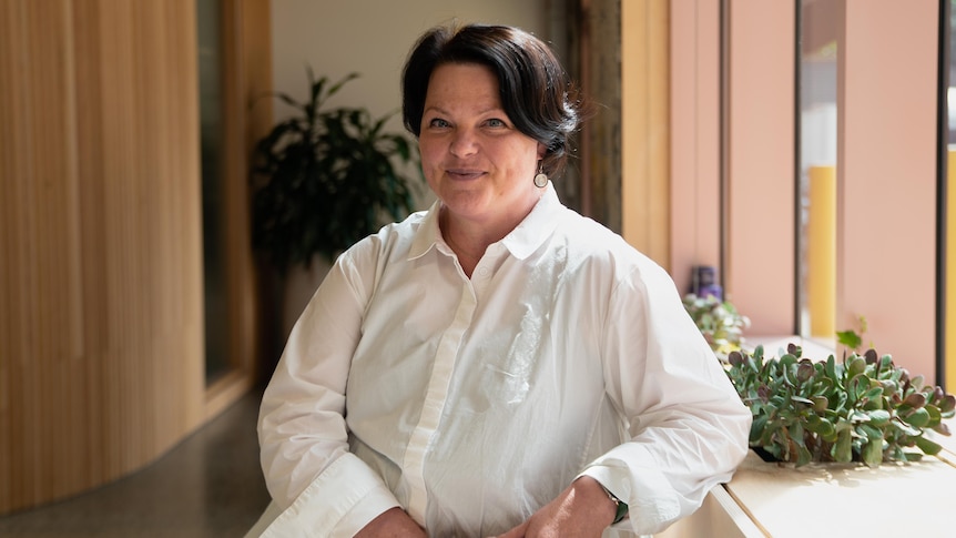 Natalie Foley, a woman who has short dark brown hair and is wearing a white shirt, poses for a photo at a sunlit bench