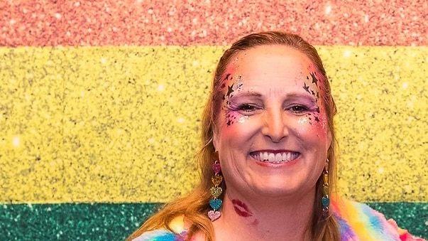 blonde middle-aged woman with rainbow stars painted on her face smiling in front of a glittery rainbow flag
