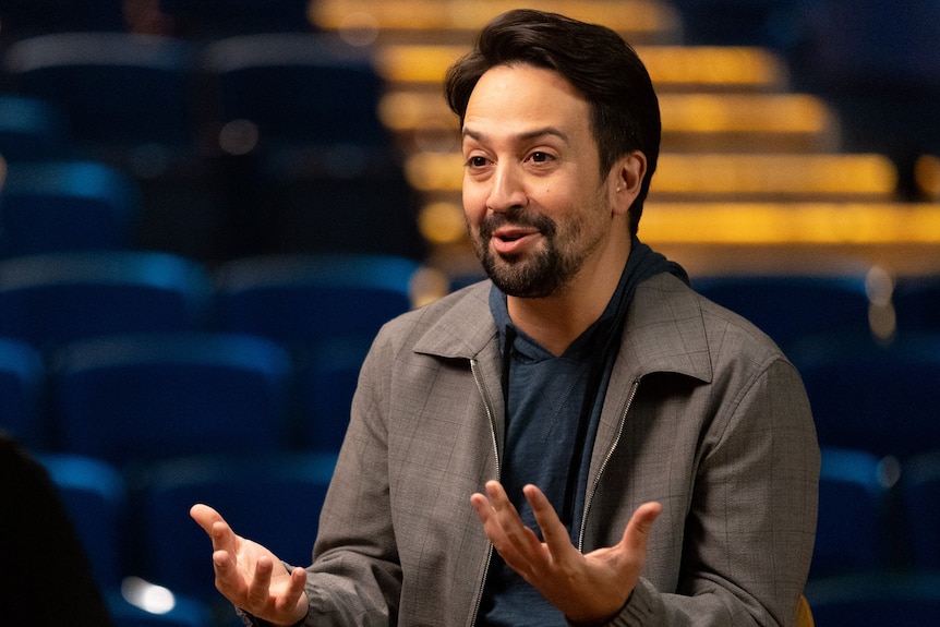 Lin-Manuel mid-sentence gesturing with his hands in front of him. He looks to the left and wears a grey jacket