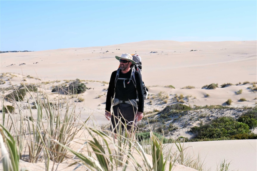 Man with hat and backpack walking in sandhills, green grass in foreground