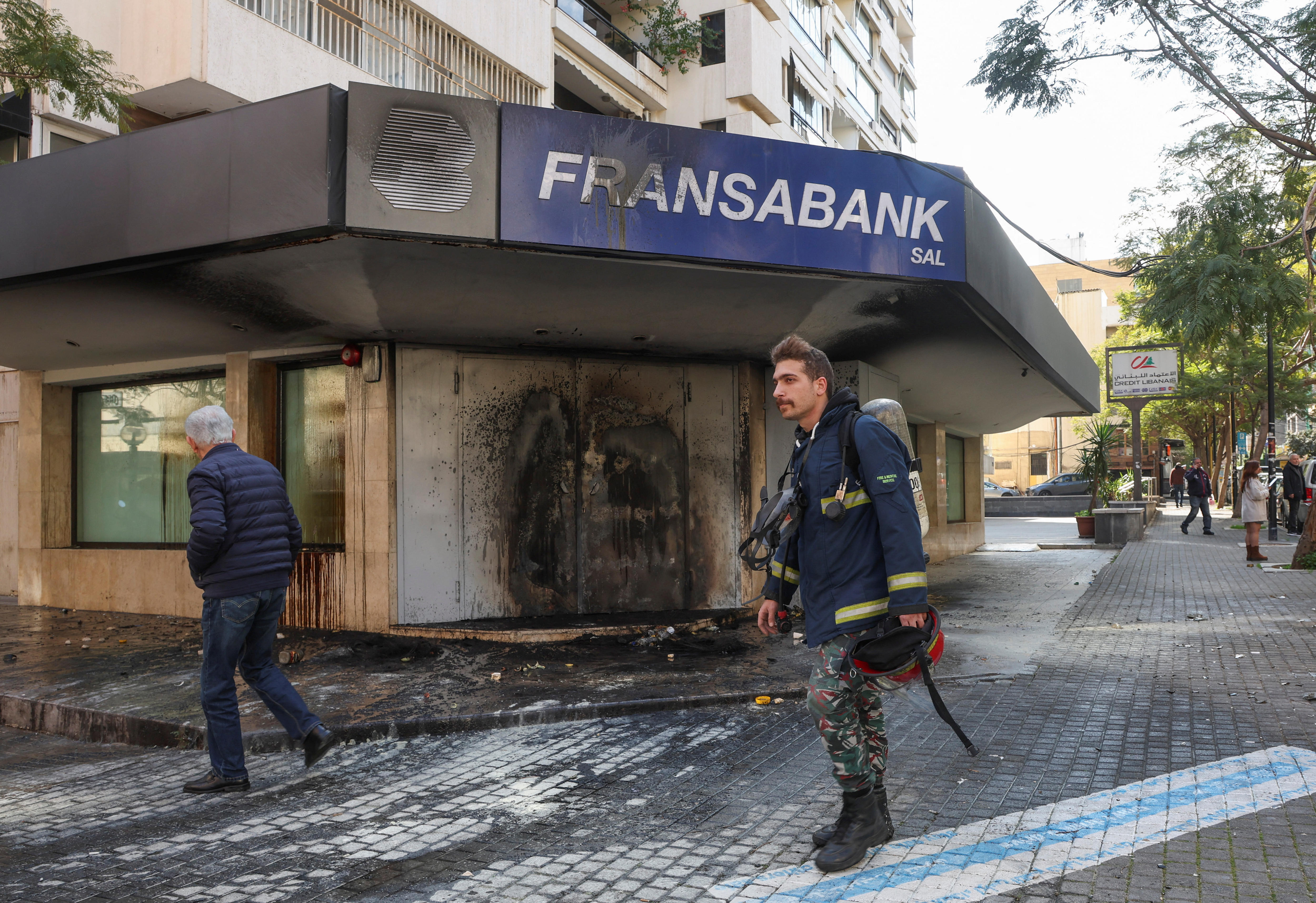 A firefighter walking past a burned building with a sign which reads "Fransabank" 