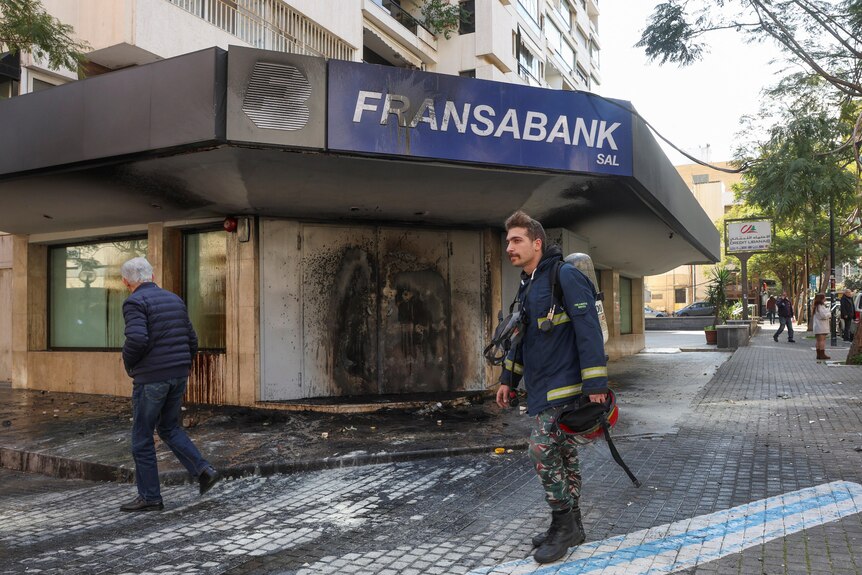 A firefighter walking past a burned building with a sign which reads "Fransabank" 