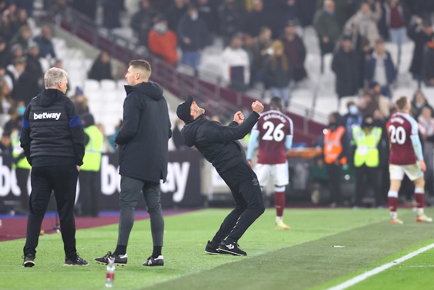 A Premier League manager stands on the sideline and throws his head back to shout joyously after his team wins.