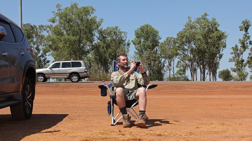 A photo of plane spotter Craig Butsch, seated on the side of a major road in Darwin with a camera in-hand.