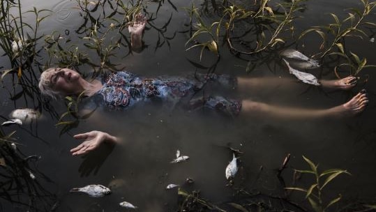 A print of a woman lying in a river surrounded by dead fish.