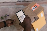 A man holds out a flat brown parcel with one hand and uses a phone to scan it with the other. Other parcels sit on the floor