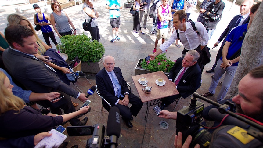 Howard and Barnett sit at a coffee table surrounded by journalists and onlookers.