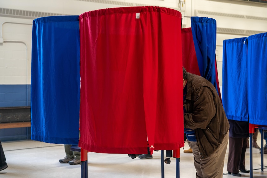 A man stands behind red and blue curtains in a circular polling booth. Similar booths are nearby.