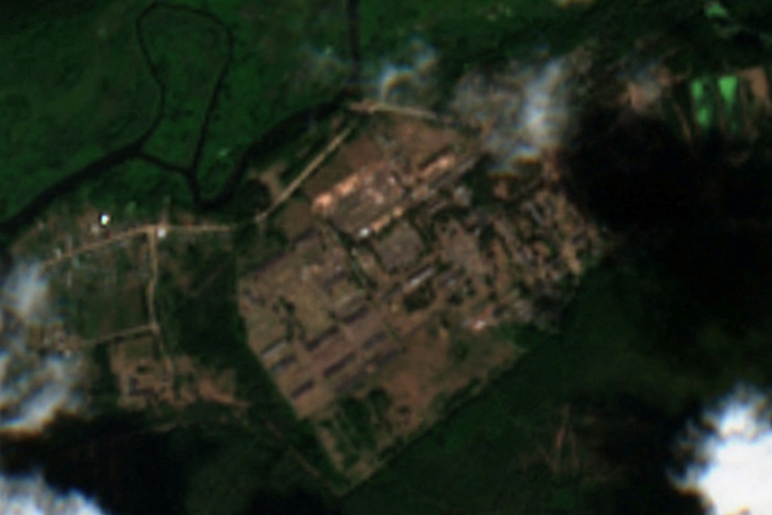 A slightly blurry satellite image shows a grid of brown structures on green land through cloud cover.