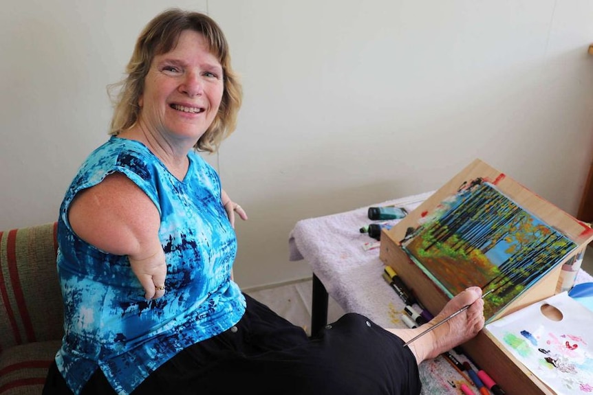 A woman holds a paintbrush between her toes. She is smiling.