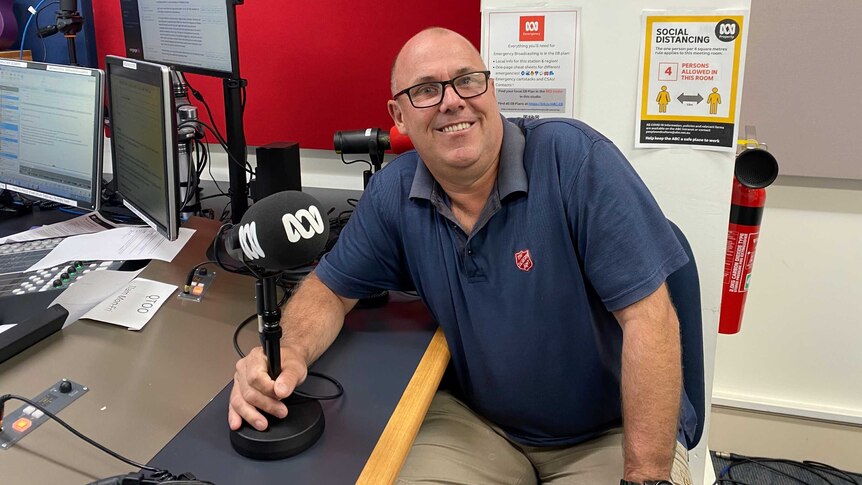 A man with glasses in a navy polo shirt sits in the ABC Wide Bay radio studio, one hand on his mic, smiling.