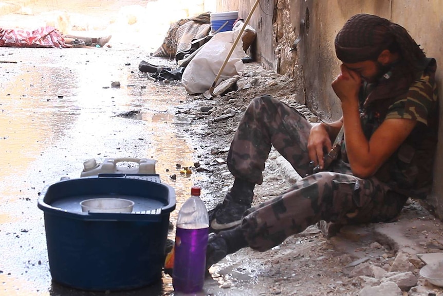 A man holds his head in his hands as a covered body can be seen in the distance.