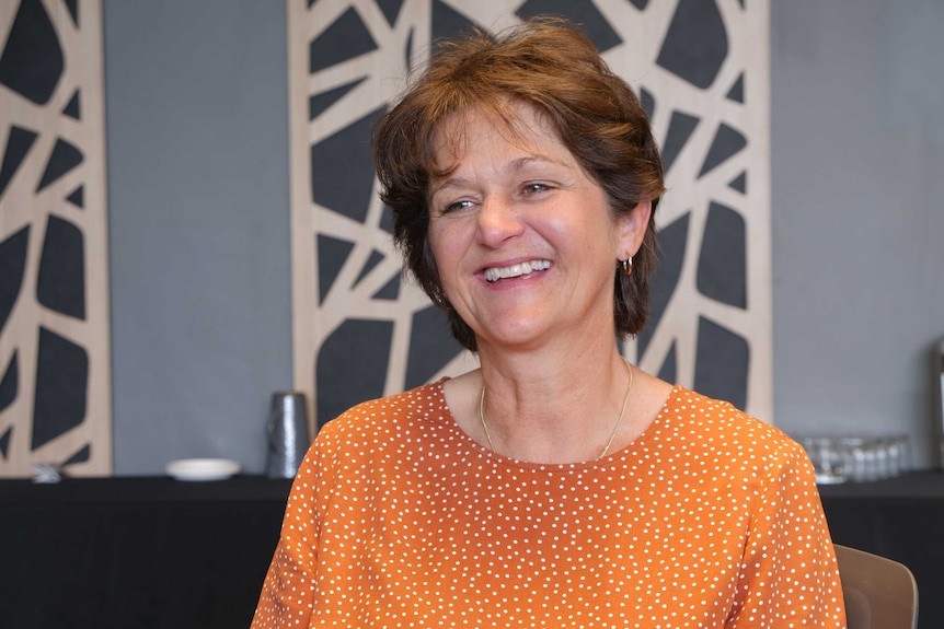 A close-up photo of a woman in her 50s smiling, sitting inside, wearing an orange spotted shirt.