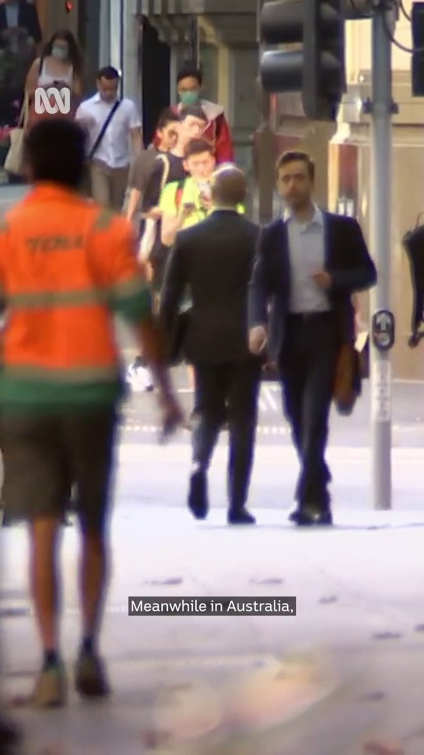 a variety of people walking through an urban environment, a traffic light is visible, somebody is wearing high-vis