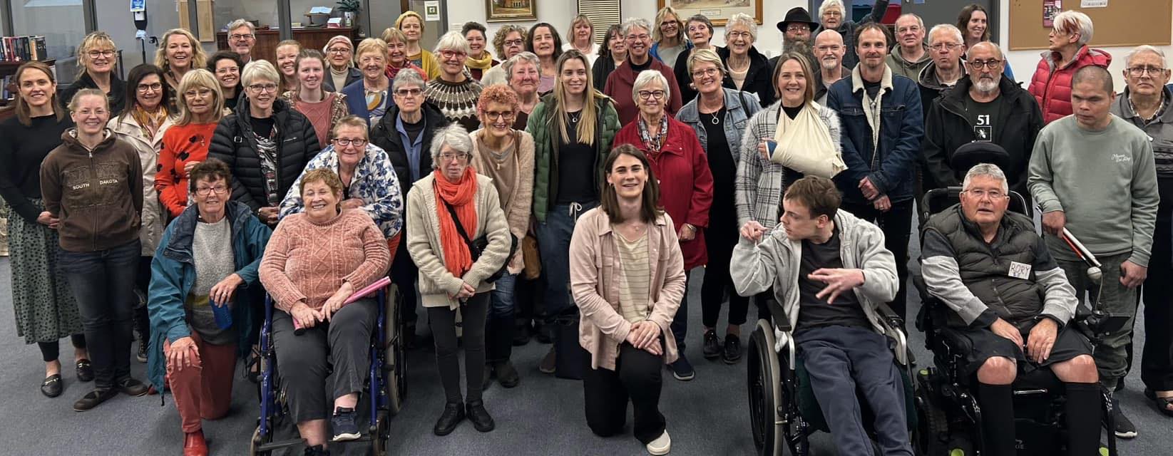 Many members of the Geelong With One Voice choir smiling at the camera and Kym Dillon kneels at the front.