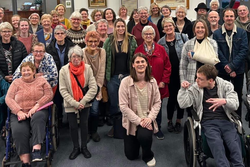 Many members of the Geelong With One Voice choir smiling at the camera and Kym Dillon kneels at the front.