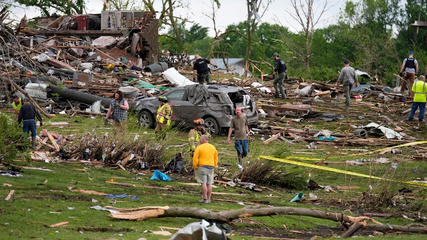A crowd of people, some in yellow reflective clothing, walk across a hillside littered with debris in the daytime.